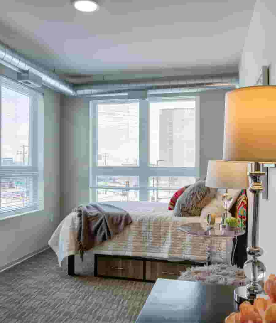 Corner bedroom with exposed HVAC ducts, bed with storage, and windows showing a city view